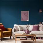 Salon lumineux et élégant avec un mur bleu pétrole, un fauteuil ocre, des coussins brique, des touches prune, des matières mates et du bois naturel.