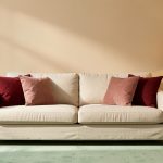 Salon lumineux aux murs beige chaud avec canapé écru, coussins rouge profond et vieux rose, tapis vert céladon et lampe jaune paille.
